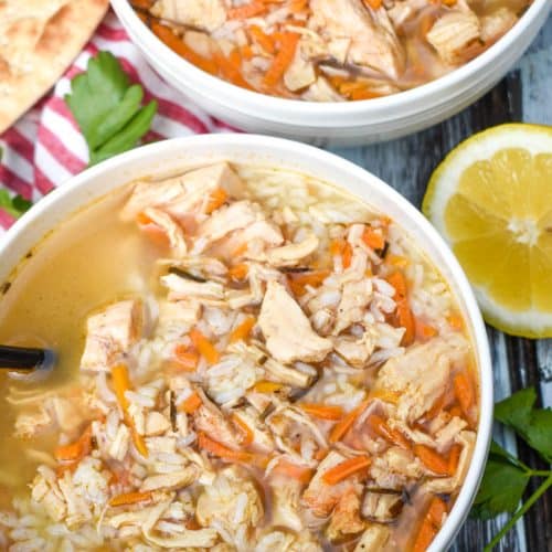 greek lemon chicken soup with rice in two white bowls with sliced lemons and fresh herbs on the side