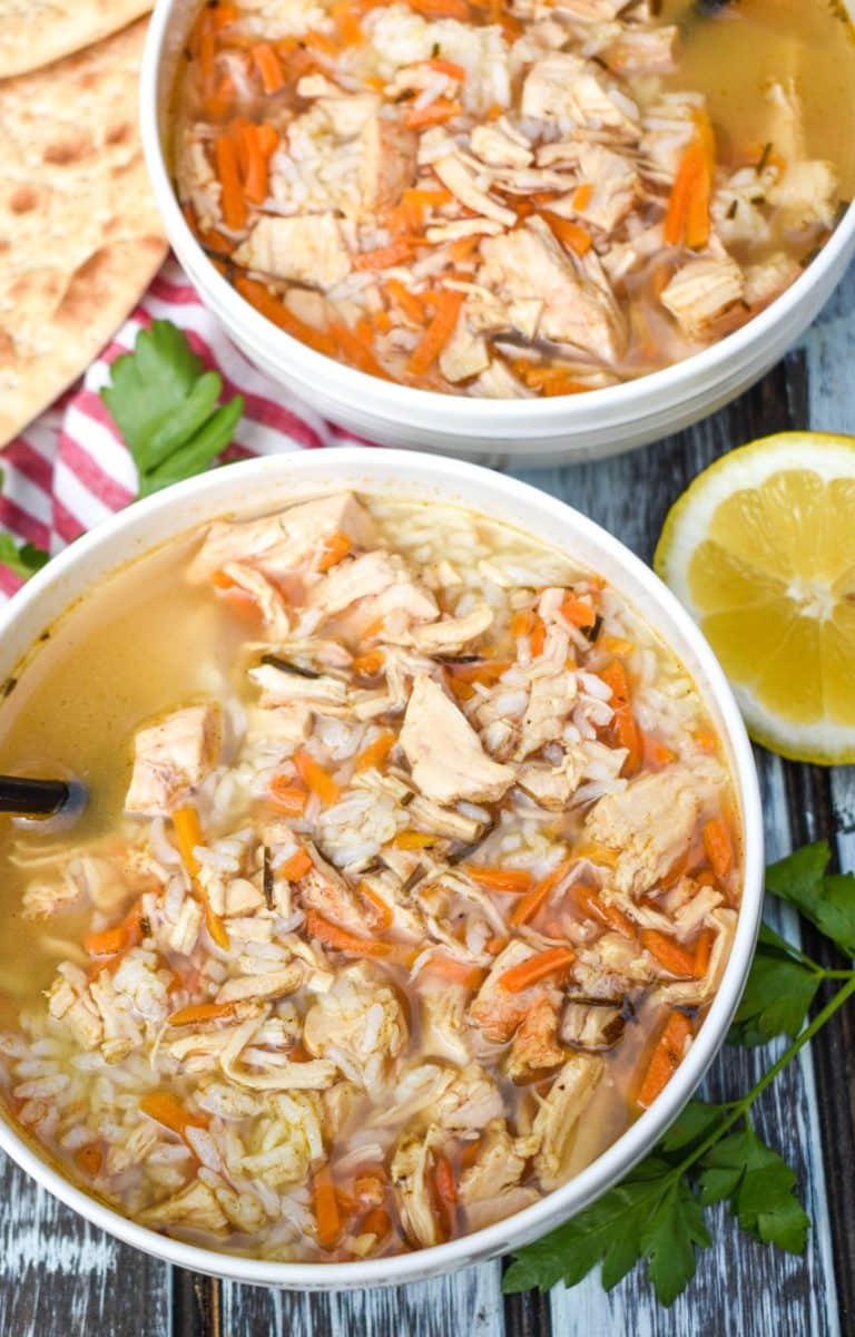 greek lemon chicken soup with rice in two white bowls with sliced lemons and fresh herbs on the side