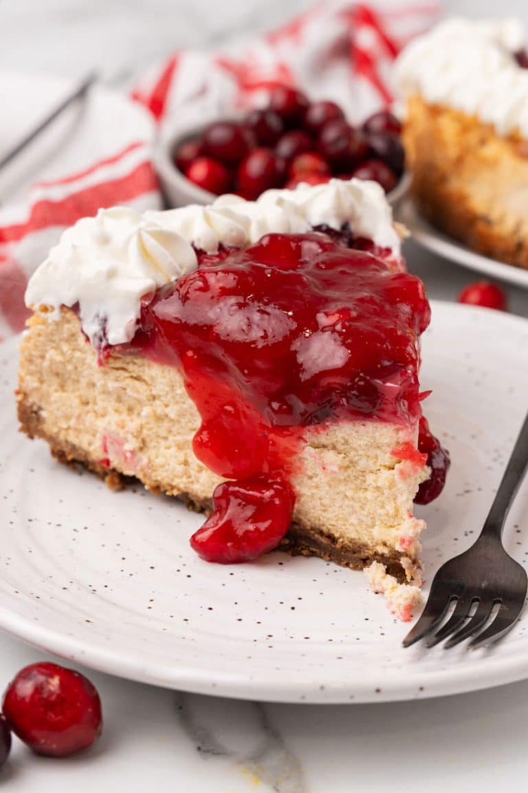 a slice of cranberry sauce topped cheesecake on a white dessert plate with a black fork resting on the edge