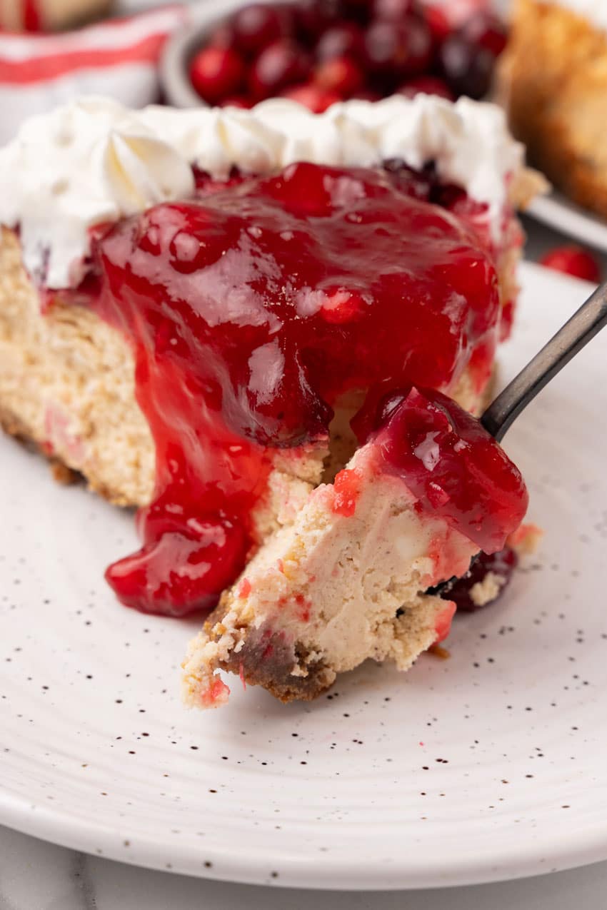 a black fork holding a scoop of eggnog cheesecake with cranberry topping resting on the edge of a white dessert plate