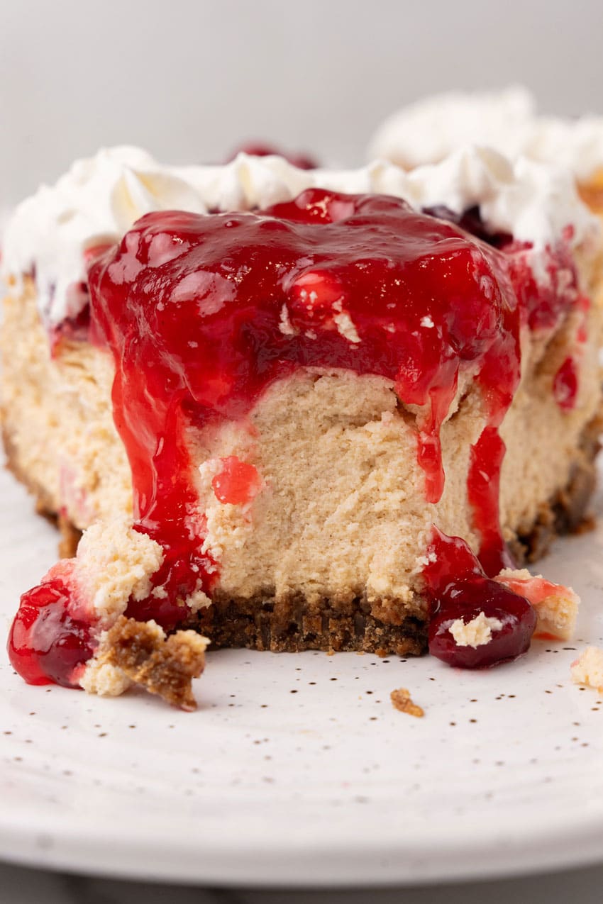 a slice of cranberry sauce topped cheesecake on a white dessert plate with a black fork resting on the edge