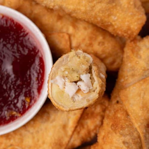 half a thanksgiving egg roll resting on a pile of fried egg rolls to reveal the filling inside with a small bowl of cranberry sauce on the side