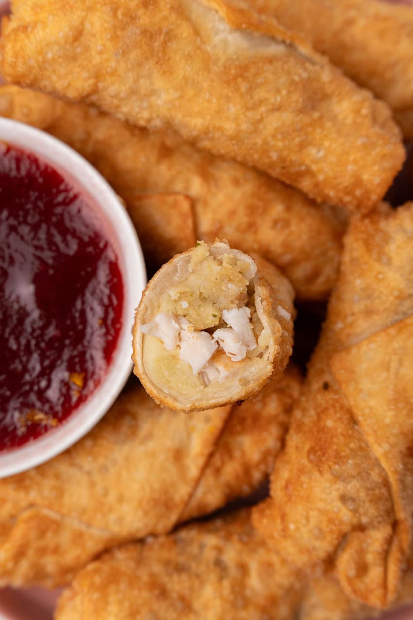 half a thanksgiving egg roll resting on a pile of fried egg rolls to reveal the filling inside with a small bowl of cranberry sauce on the side