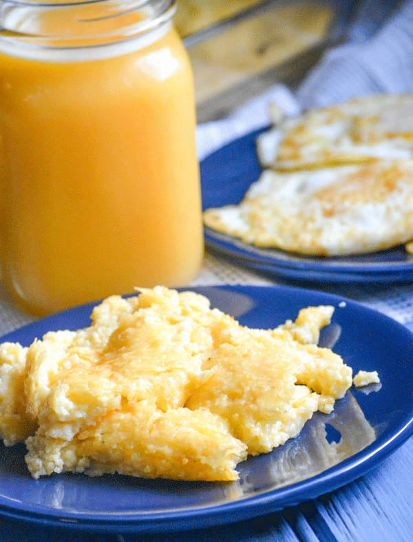 A SCOOP OF THREE CHEESE GRITS CASSEROLE ON A BLUE PLATE WITH A GLASS OF ORANGE JUICE IN THE BACKGROUND