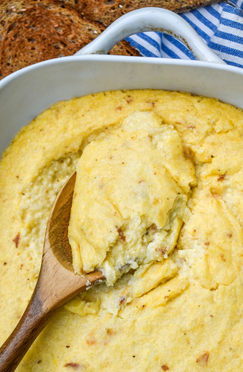 a wooden spoon scooping cheese grits out of a white casserole dish