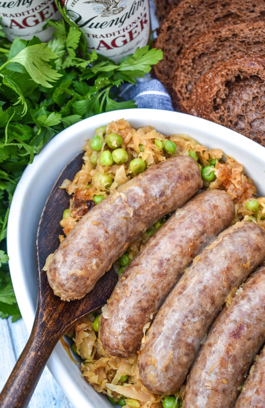 A WOODEN SPATULA SCOOPING A COOKED BEER BRATWURST AND SAUERKRAUT OUT OF A WHITE SERVING DISH