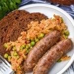 BEER BRATWURST WITH SAUERKRAUT ON A WHITE PLATE WITH TWO THICK SLICES OF PUMPERNICKLE BREAD AND A SILVER FORK RESTING ON THE EDGE