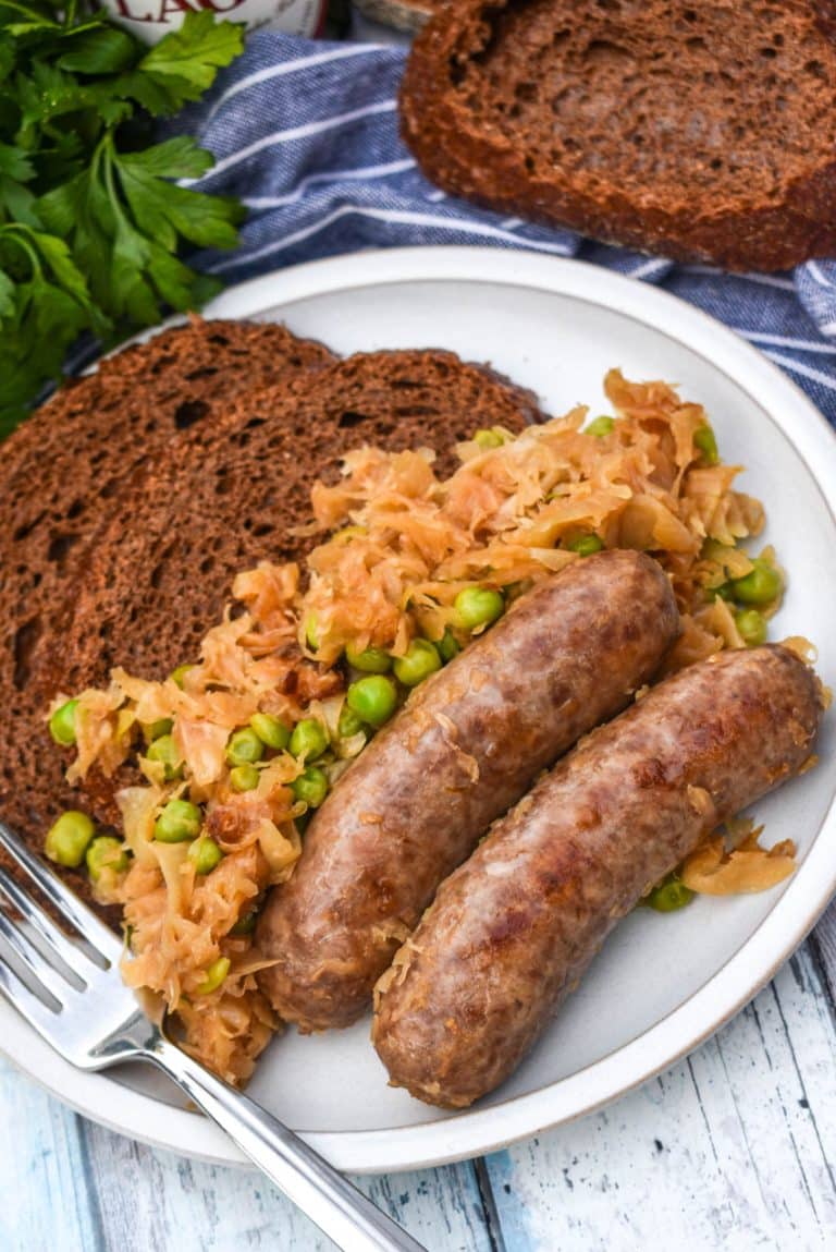 BEER BRATWURST WITH SAUERKRAUT ON A WHITE PLATE WITH TWO THICK SLICES OF PUMPERNICKLE BREAD AND A SILVER FORK RESTING ON THE EDGE