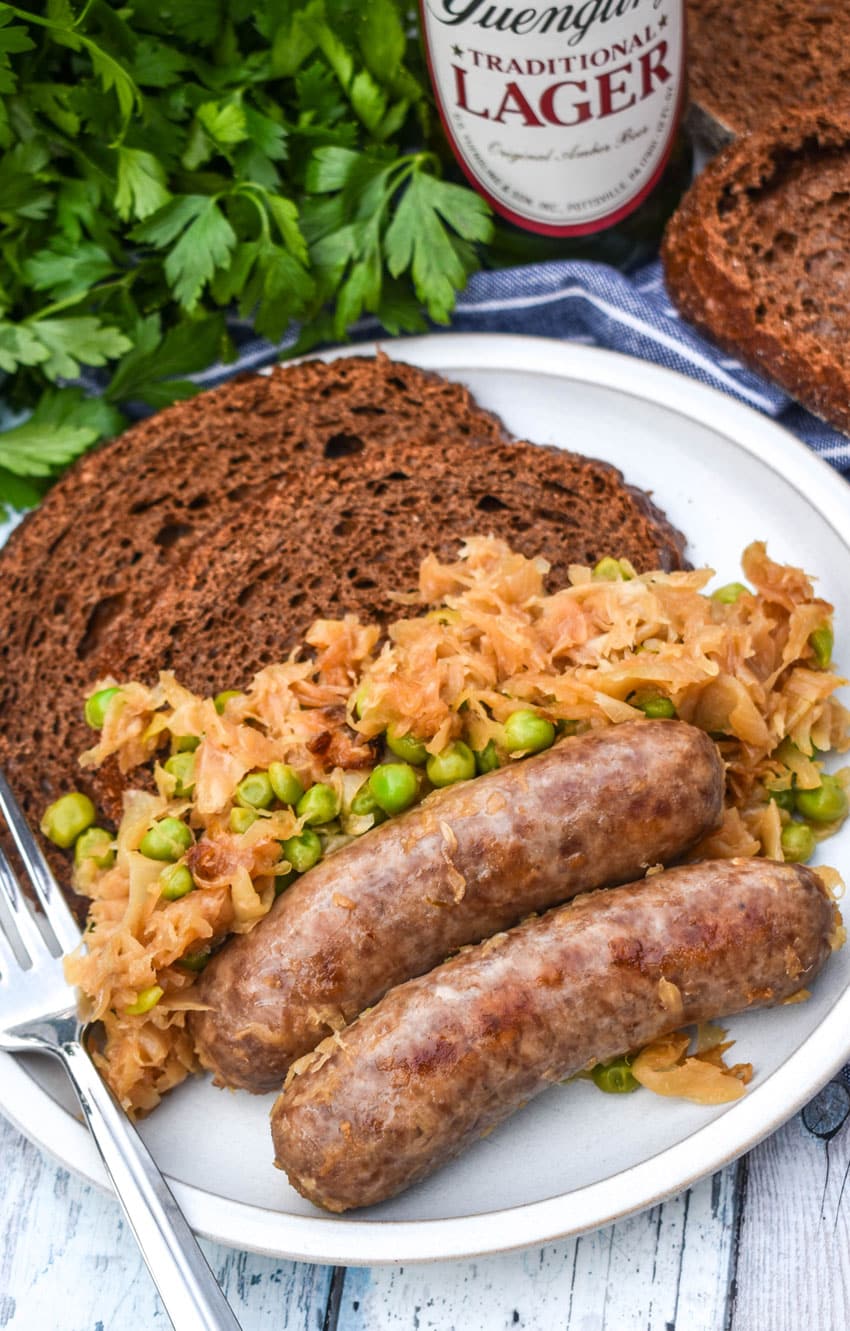 BEER BRATWURST WITH SAUERKRAUT ON A WHITE PLATE WITH TWO THICK SLICES OF PUMPERNICKLE BREAD AND A SILVER FORK RESTING ON THE EDGE