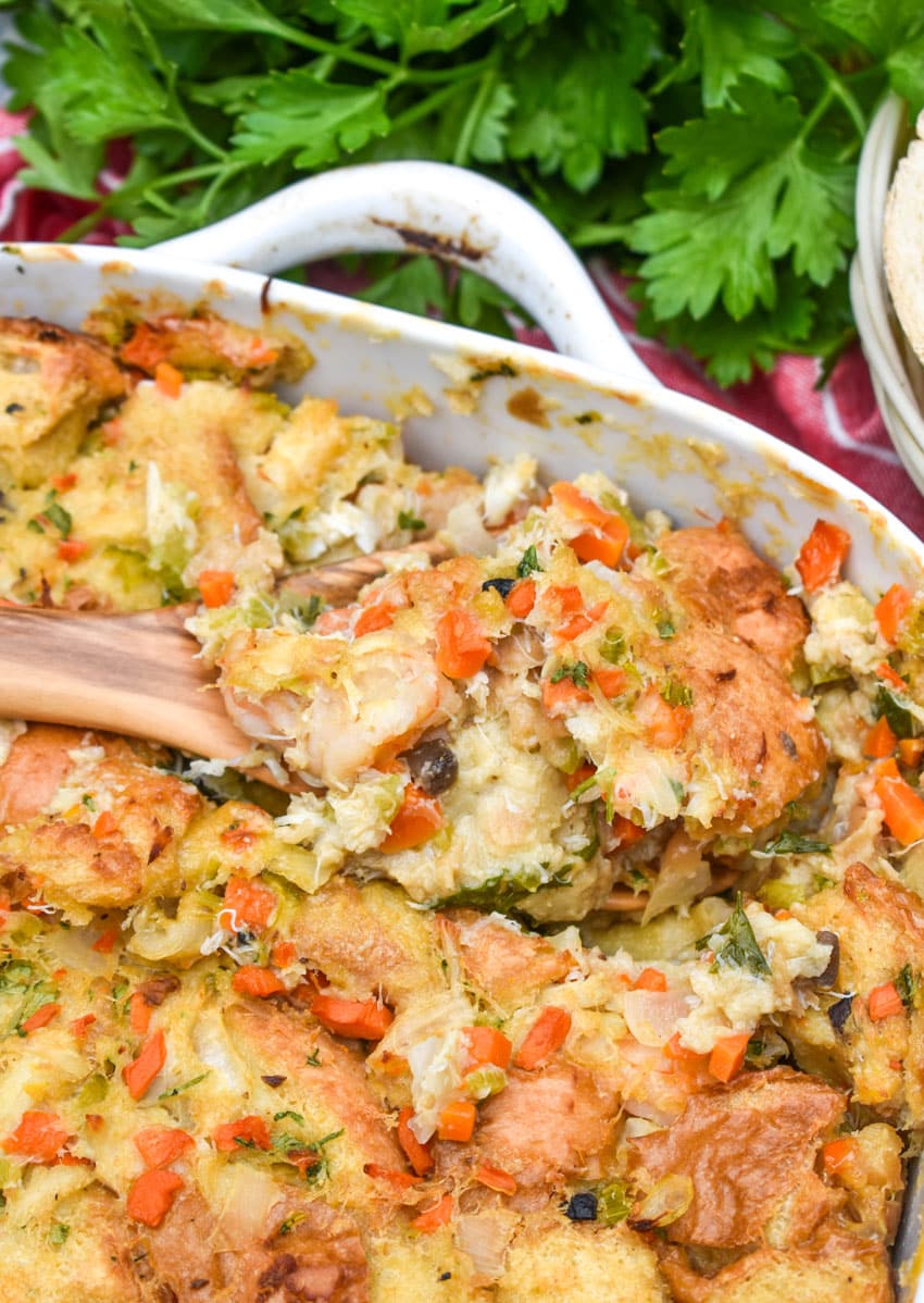 a wooden spoon scooping baked seafood stuffing out of a white casserole dish