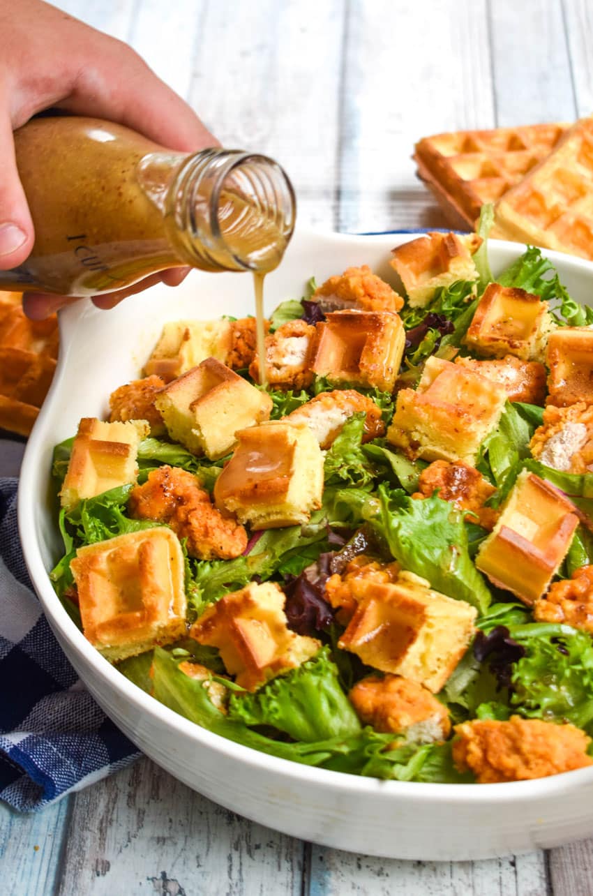 maple dijon vinaigrette being poured from a glass jar over chicken and waffle salad in a white serving bowl