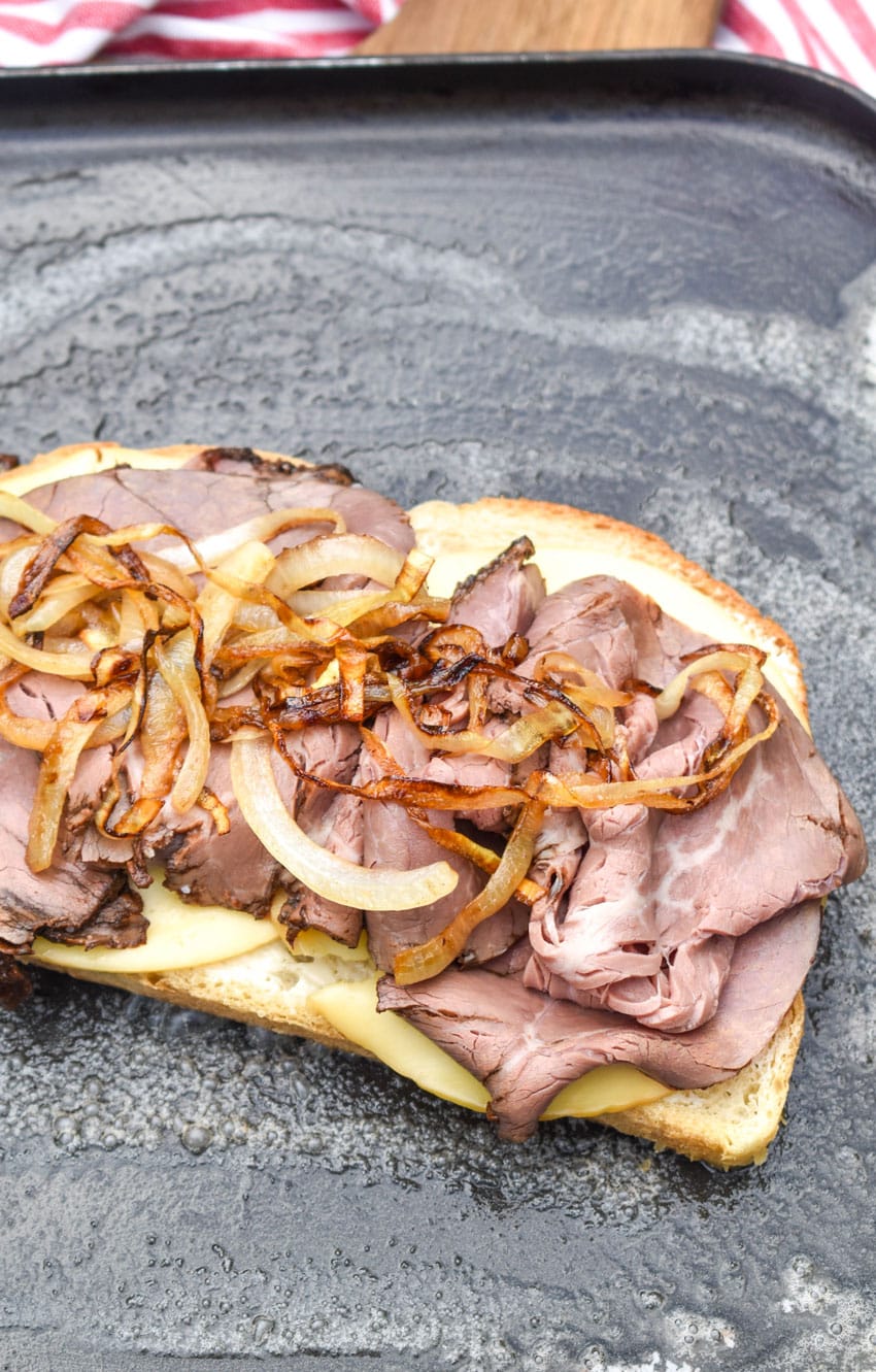 a slice of sourdough bread topped with sliced gouda cheese, roast beef, and caramelized onions on a buttered skillet