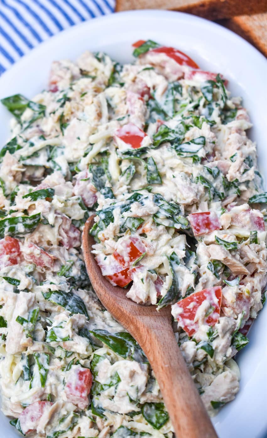 a wooden spoon scooping spinach and tomato tuna salad out of a white bowl