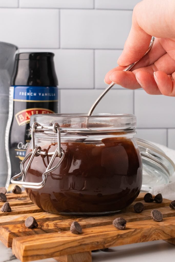 a hand holding a silver spoon in a glass jar filled with bailey's chocolate sauce