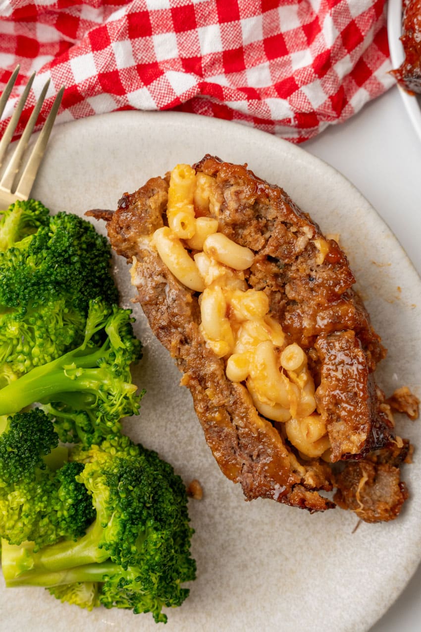 a slice of mac and cheese stuffed meatloaf with steamed broccoli on the side of a white dinner plate
