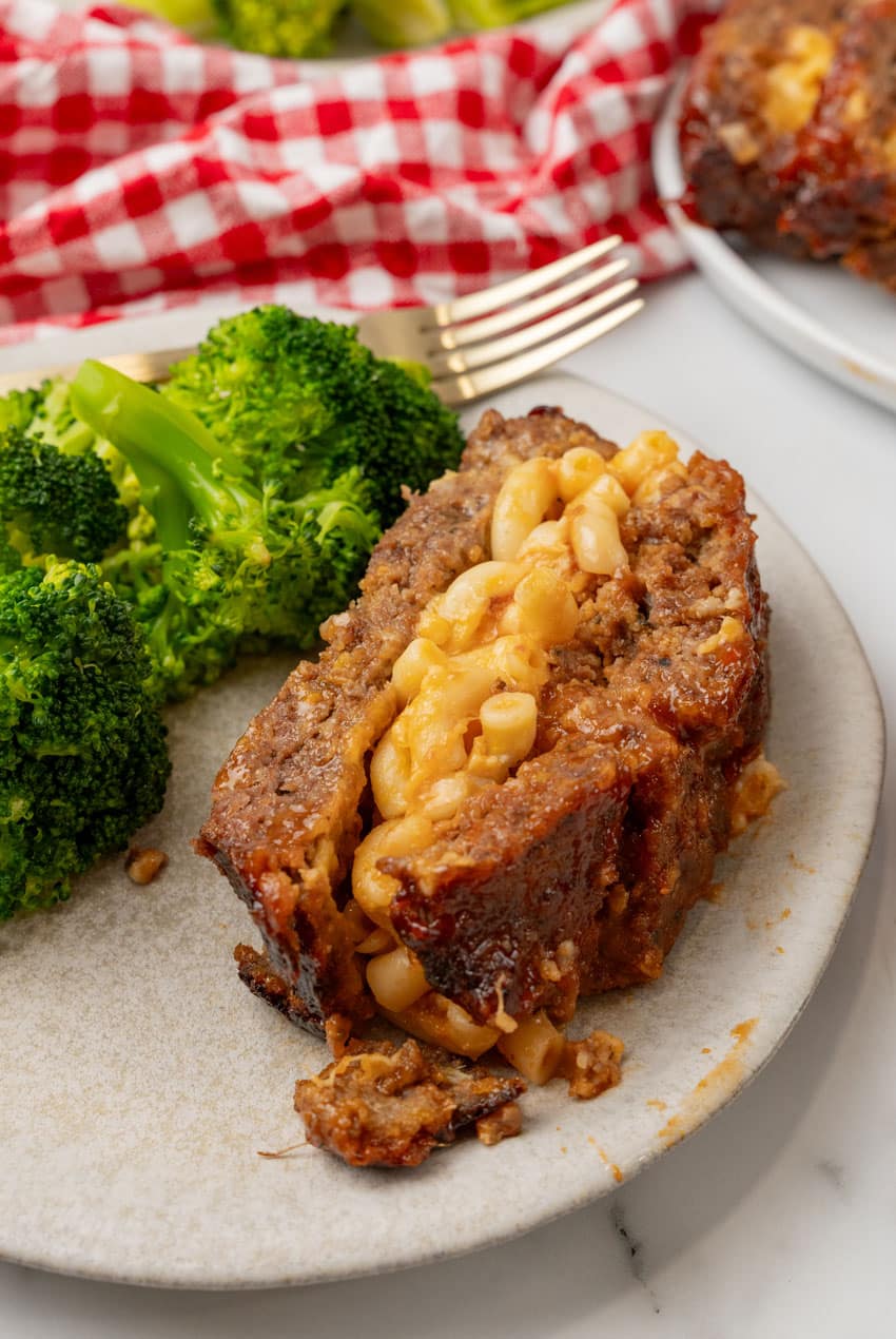 a slice of mac and cheese stuffed meatloaf with steamed broccoli on the side of a white dinner plate
