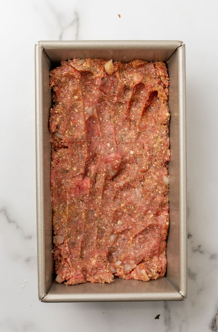 seasoned ground beef meatloaf mixture spread out in an even layer in a metal loaf pan