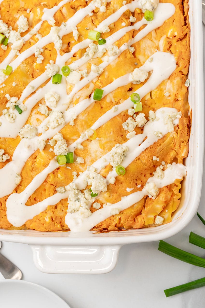 cheesy buffalo chicken lasagna in a white baking dish topped with a drizzle of ranch dressing, crumbled cheese, and sliced green onions