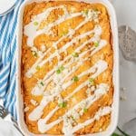 cheesy buffalo chicken lasagna in a white baking dish topped with a drizzle of ranch dressing, crumbled cheese, and sliced green onions