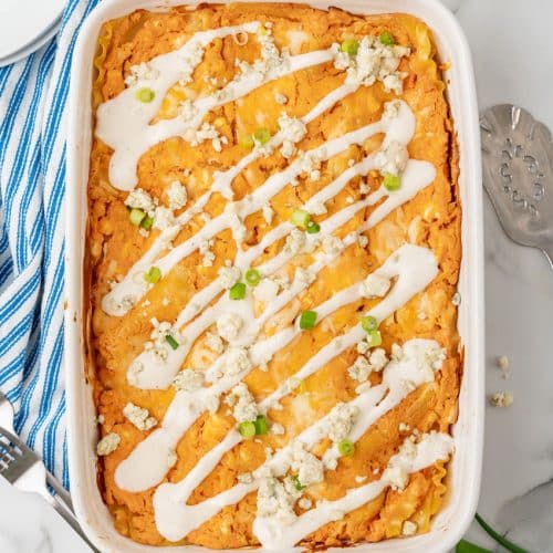 cheesy buffalo chicken lasagna in a white baking dish topped with a drizzle of ranch dressing, crumbled cheese, and sliced green onions
