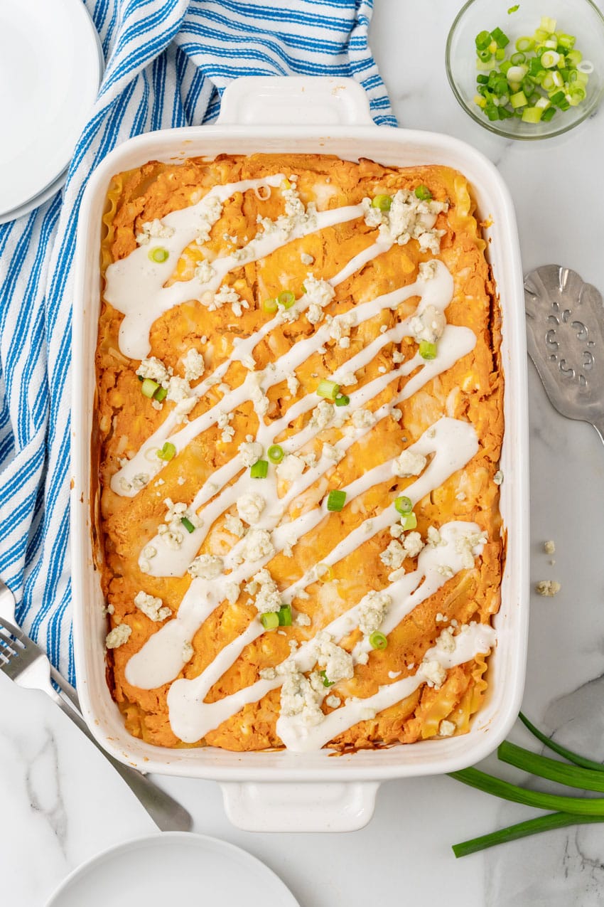 cheesy buffalo chicken lasagna in a white baking dish topped with a drizzle of ranch dressing, crumbled cheese, and sliced green onions