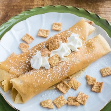 TWO CINNAMON TOAST CRUNCH CREPES TOPPED WITH WHIPPED CREAM AND CEREAL PIECES ON A WHITE PLATE