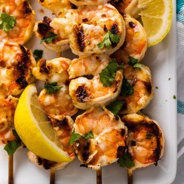 grilled nam prik shrimp skewers stacked on a white square plate with fresh lemon wedges