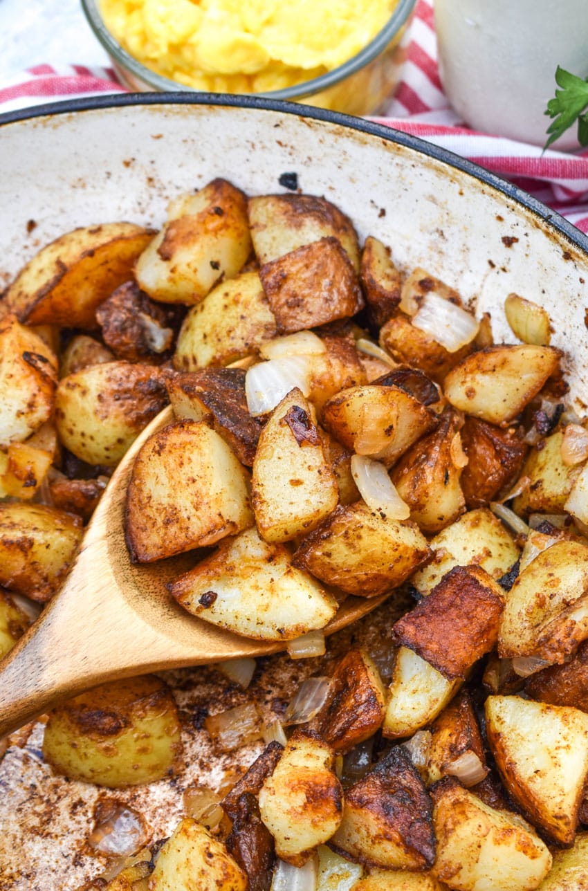 a wooden spoon scooping home fried breakfast potato recipe out of a cast iron skillet