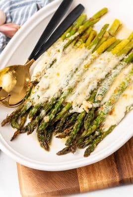 cheesy garlic roasted asparagus on a white serving platter resting on a wooden cutting board