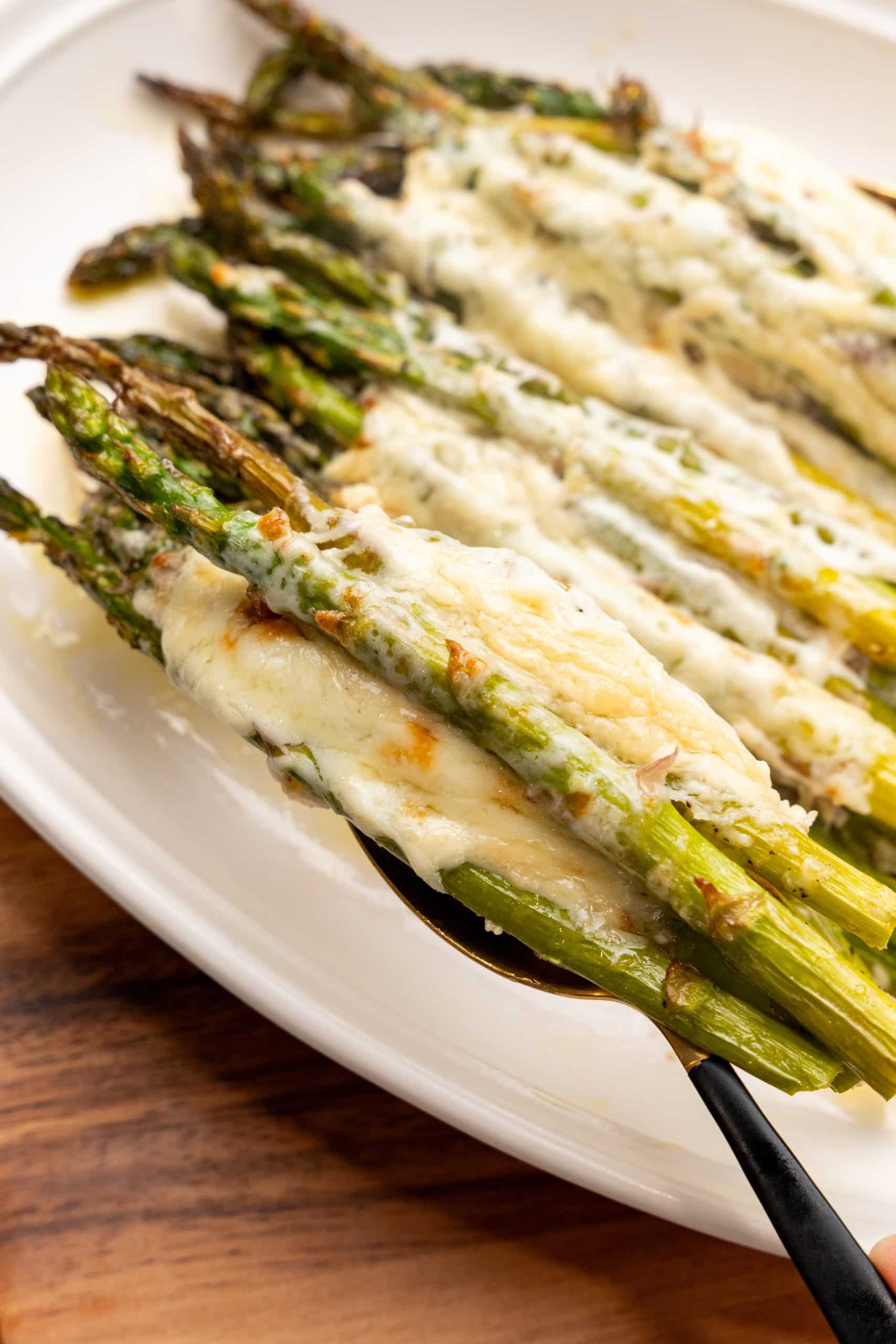a spoon lifting cheesy garlic roasted asparagus off a white serving platter