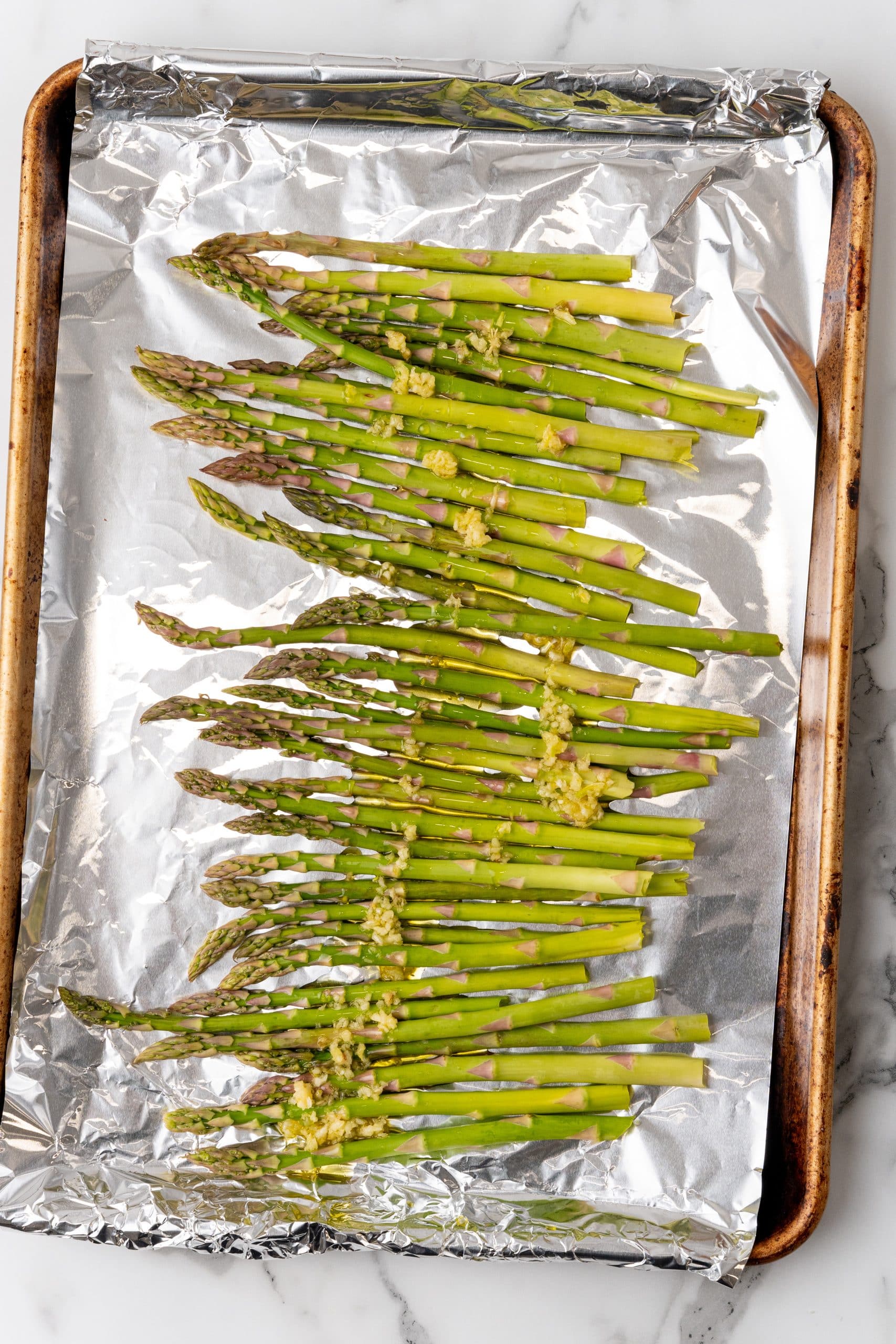 stalks of asparagus topped with minced garlic arranged in a single row on a foil lined sheet pan