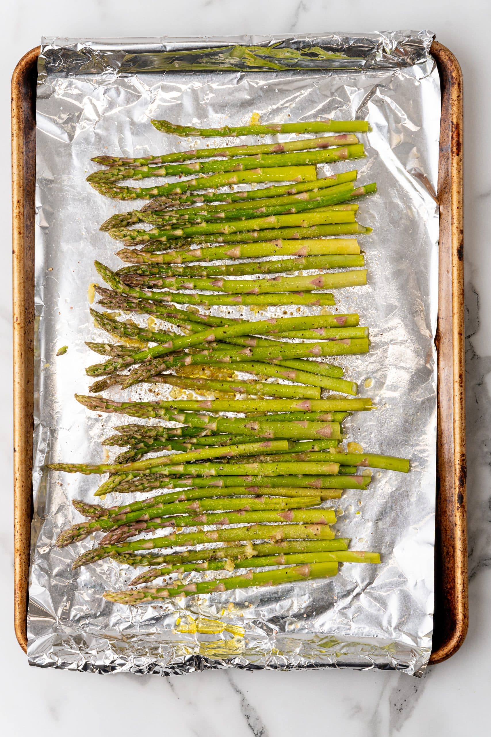 garlic and olive oil coated stalks of asparagus arranged in a single row on a foil lined baking sheet