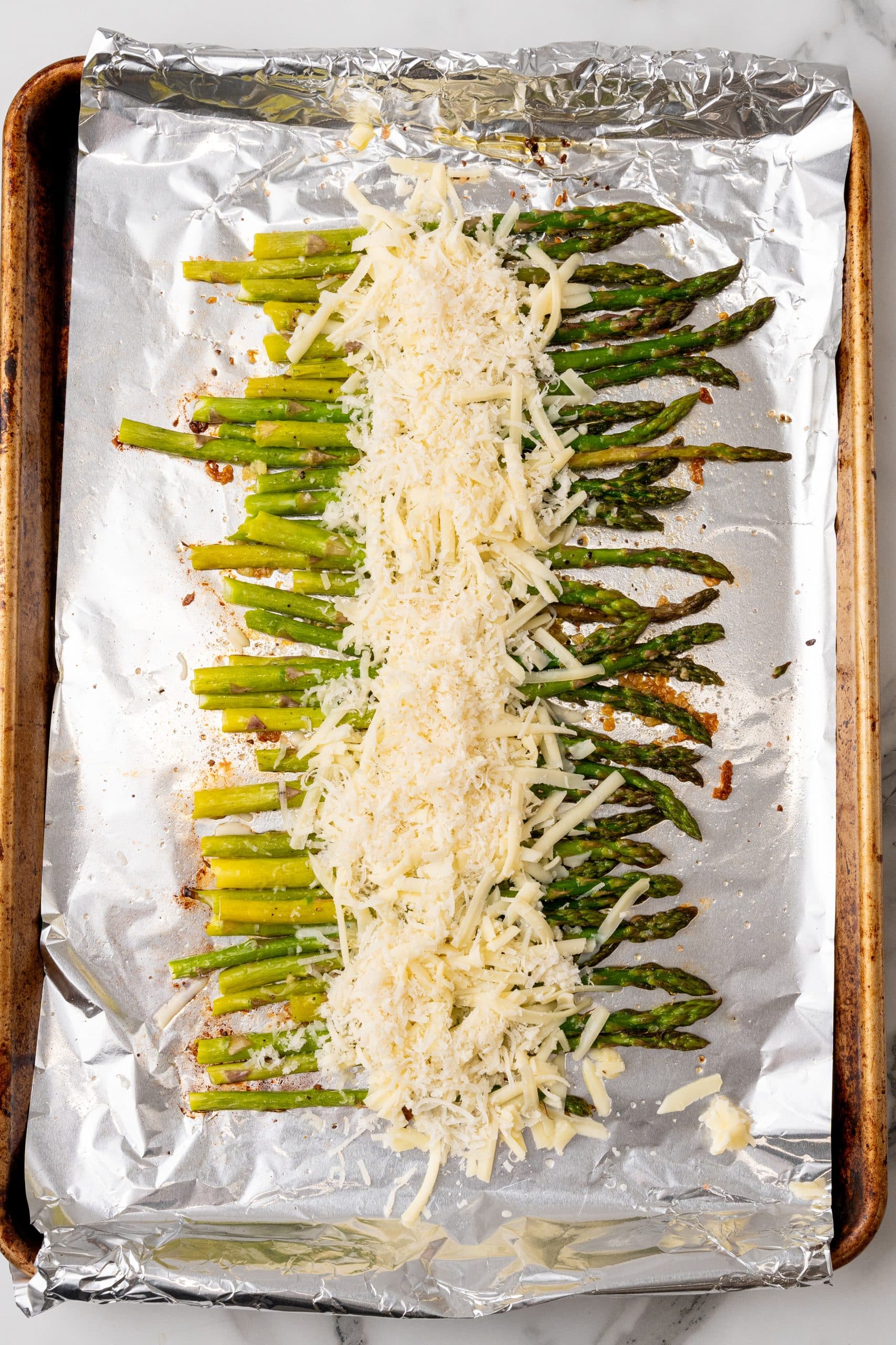 a row of garlic roasted asparagus topped with shredded cheese on a foil lined sheet pan