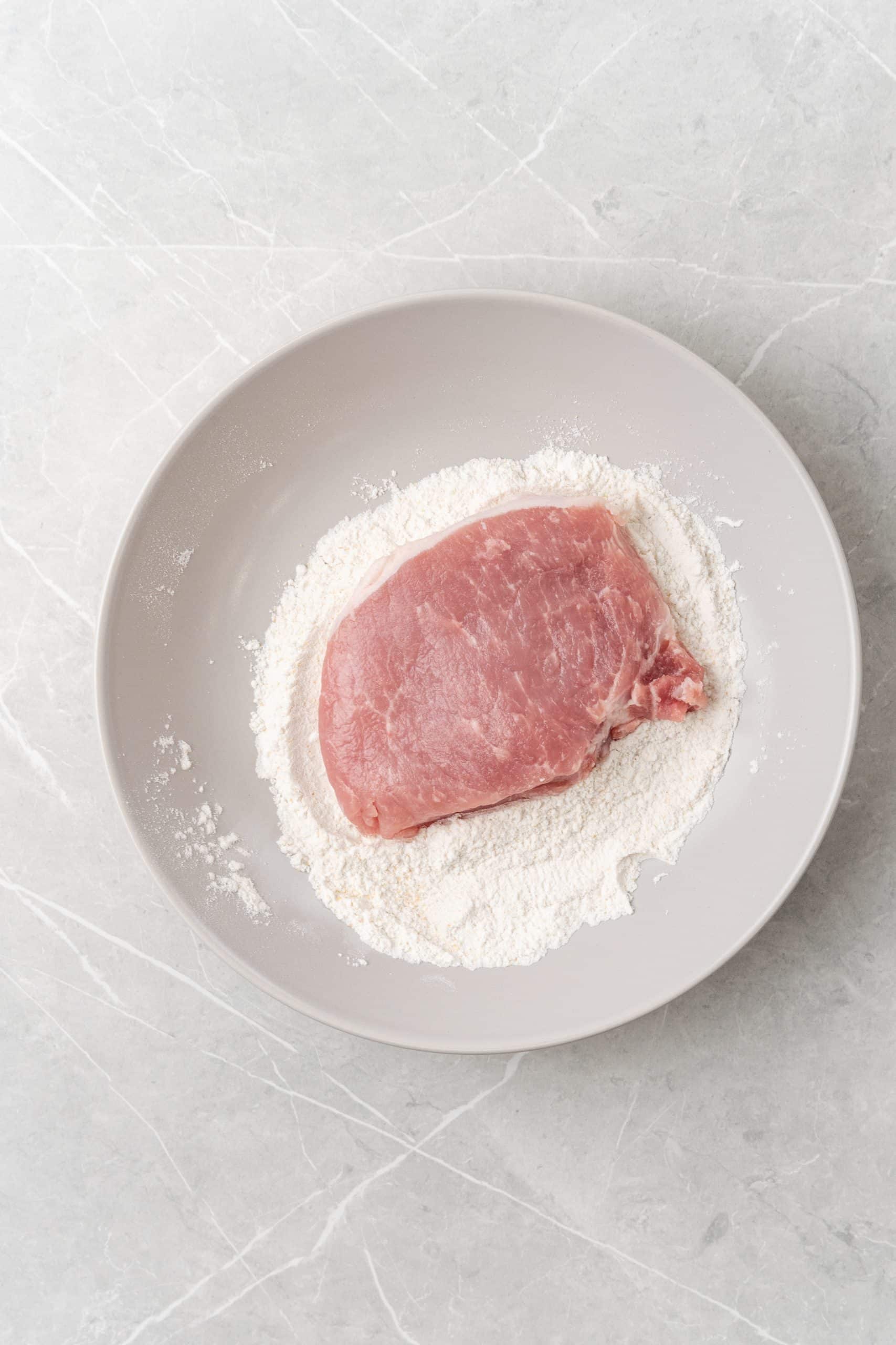 a pork chop resting on top of seasoned flour in a white mixing bowl