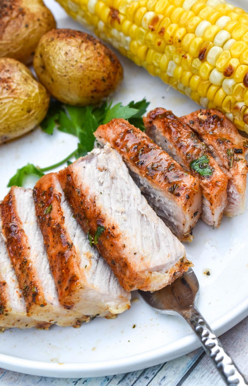 a sliced ranch pork chop topped on a white plate with potatoes and corn on the cob on the side