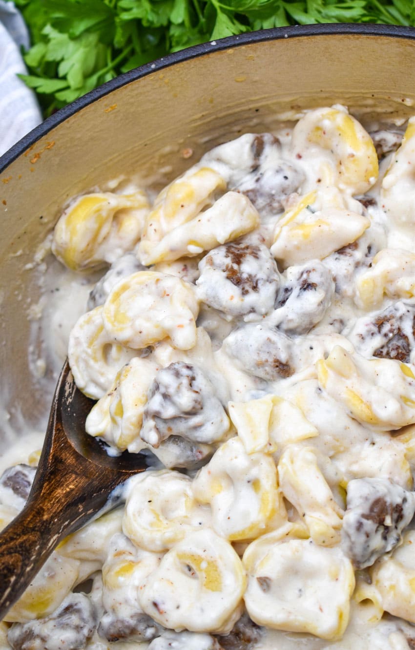 a wooden spoon scooping sausage tortellini alfredo out of a large red dtuch oven