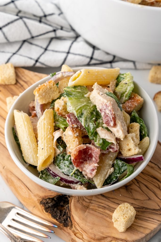 chicken caesar pasta salad in a small white bowl on a wooden cutting board