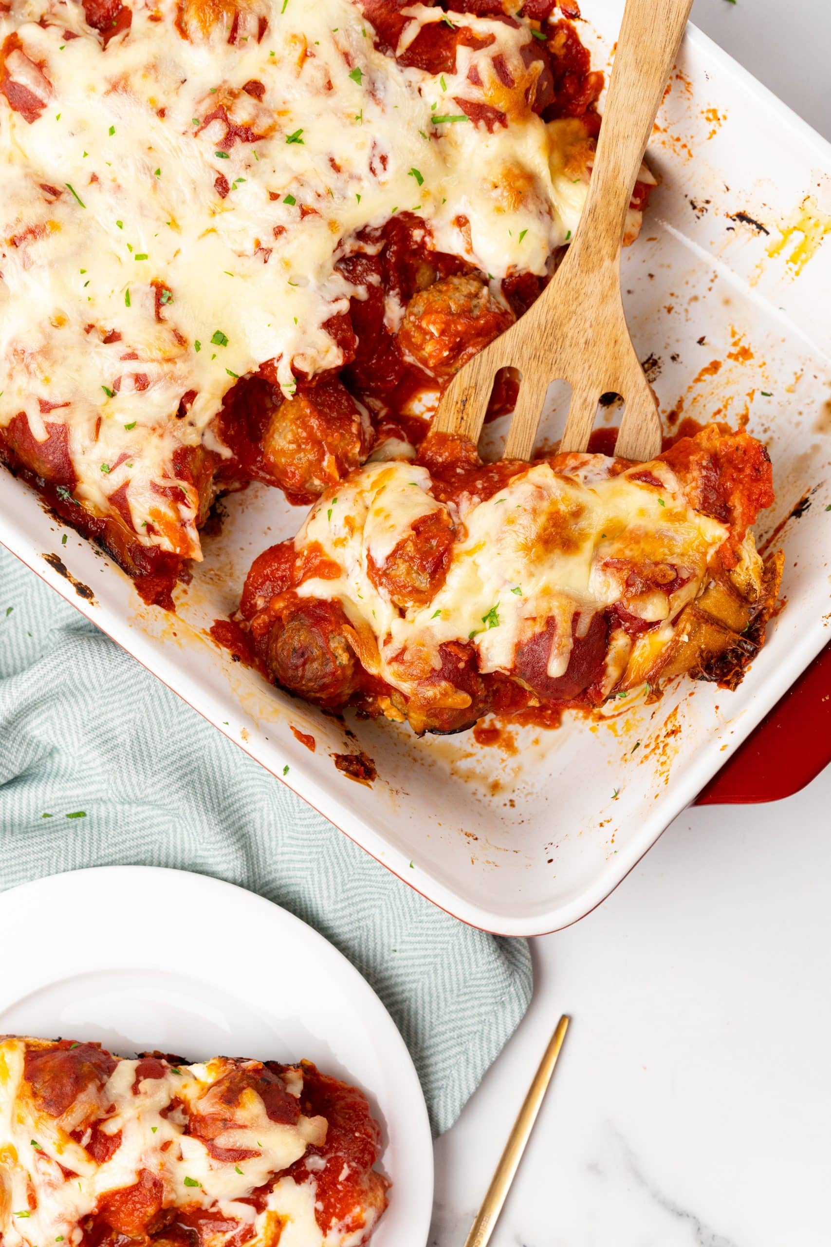 a wooden spatula scooping a slice of meatball sub casserole out of a white baking dish