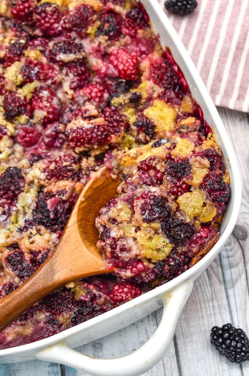 a wooden spoon digging into a dump and bake blackberry cobbler in a white casserole dish
