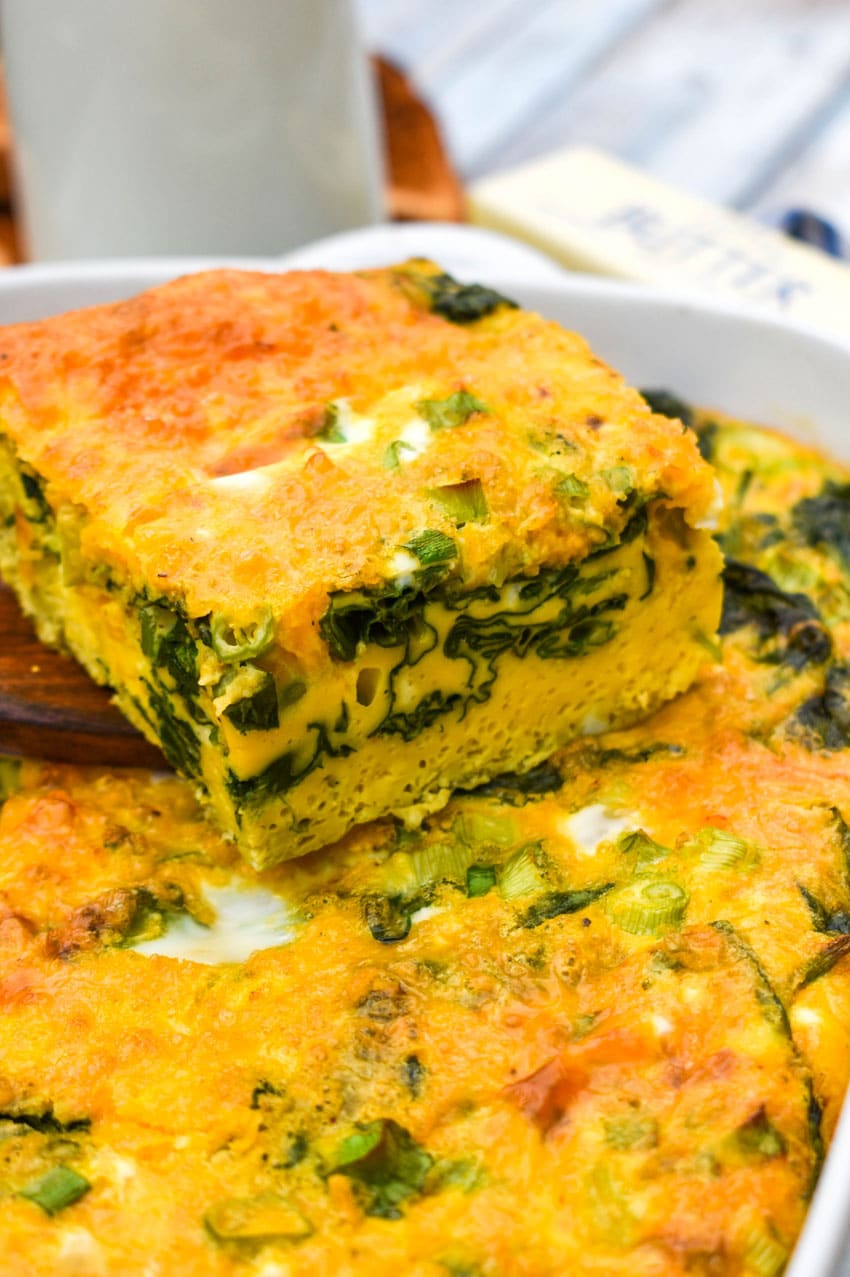 a wooden spatula holding up a slice of cheesy egg casserole with spinach