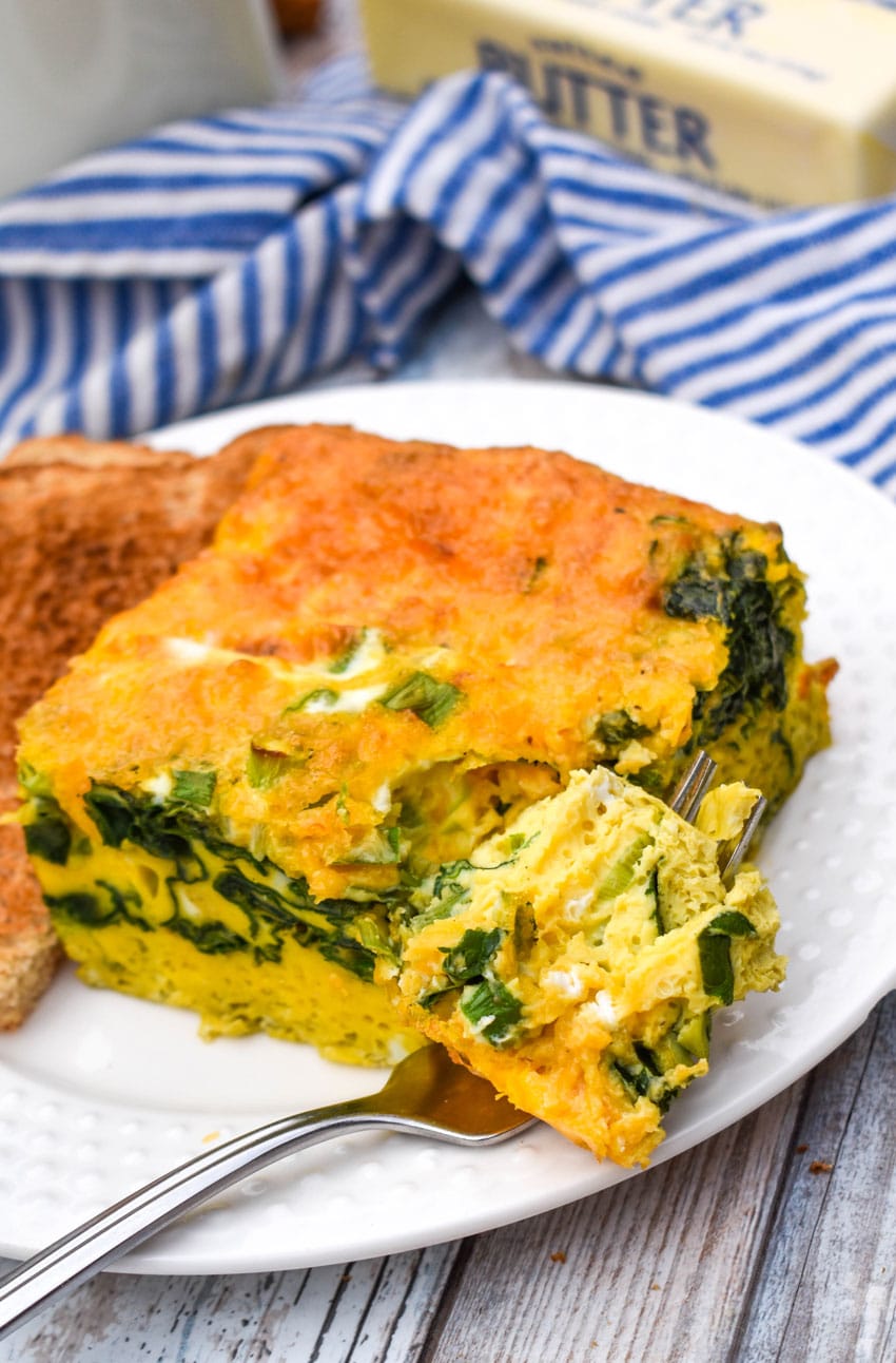 a silver fork resting on a white plate with a slice of cheesy egg casserole with spinach