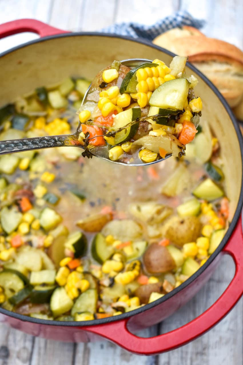 a silver ladle lifting a scoop of summer soup with zucchini and corn out of a red pot