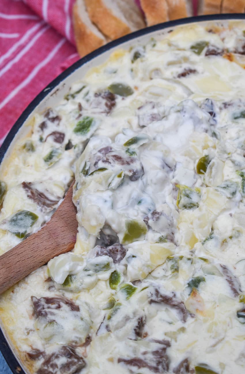 a wooden spoon scooping baked philly cheesesteak dip out of a cast iron skillet