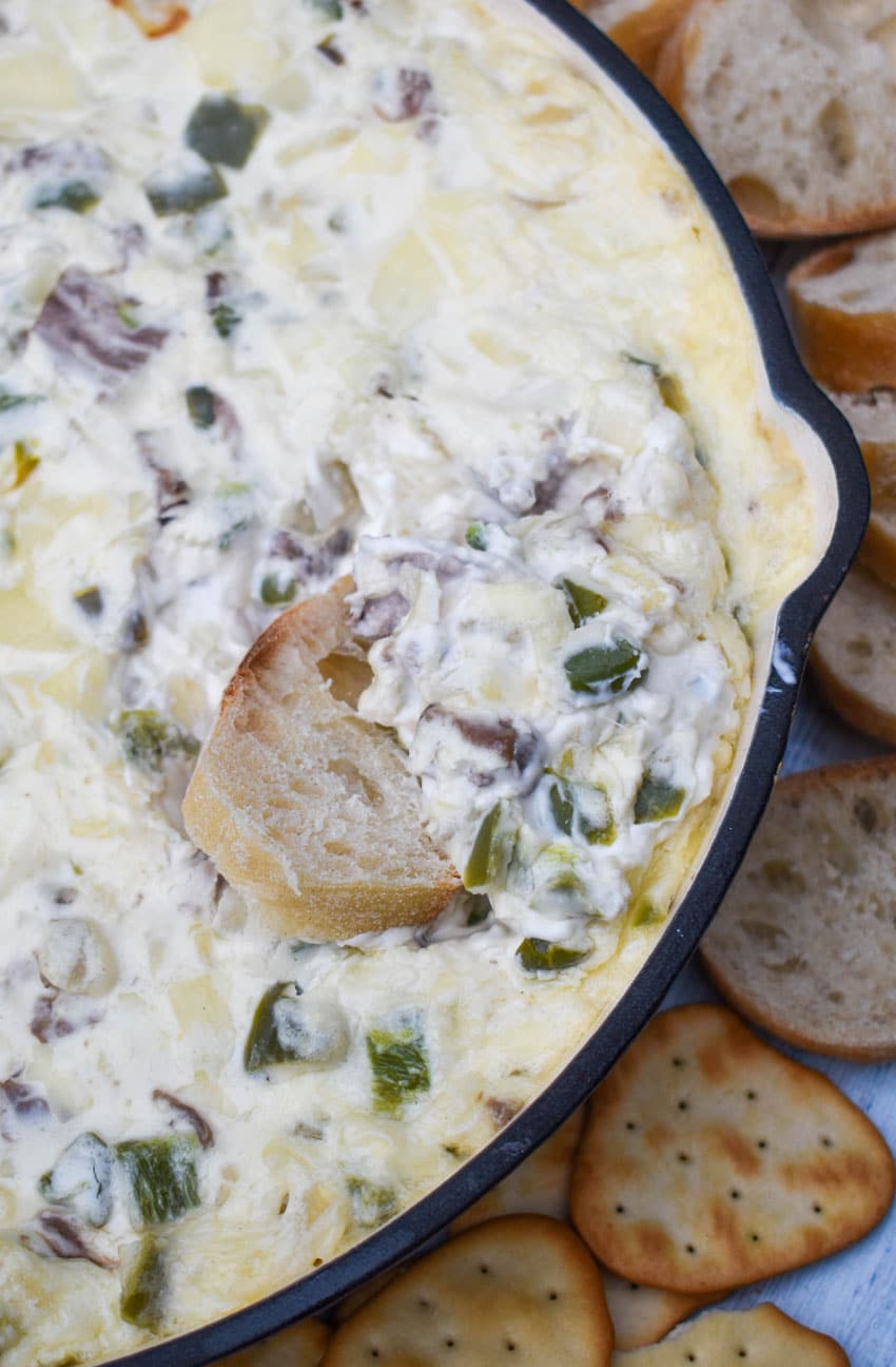 a slice of baguette bread scooping baked philly cheesesteak dip out of a cast iron skillet