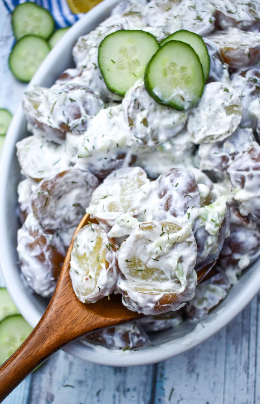a wooden spoon scooping tzatziki potato salad out of a white serving dish