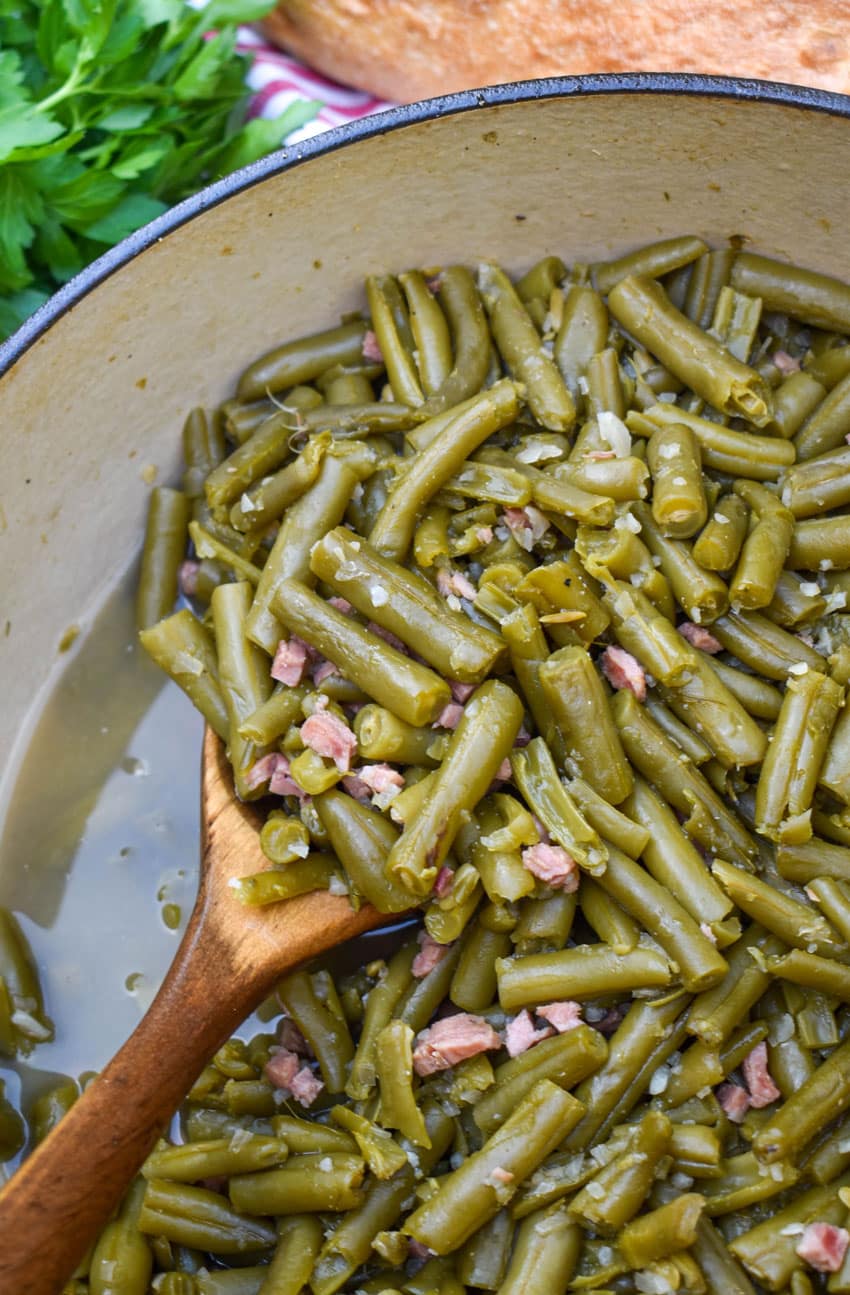 a wooden spoon scooping a canned green beans recipe out of a red dutch oven