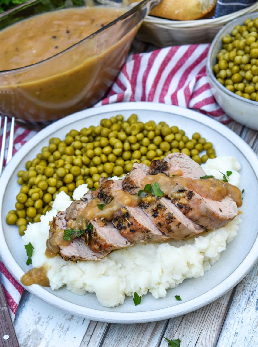 slices of pork tenderloin with apple cider gravy over mashed potatoes next to baby peas on a white plate