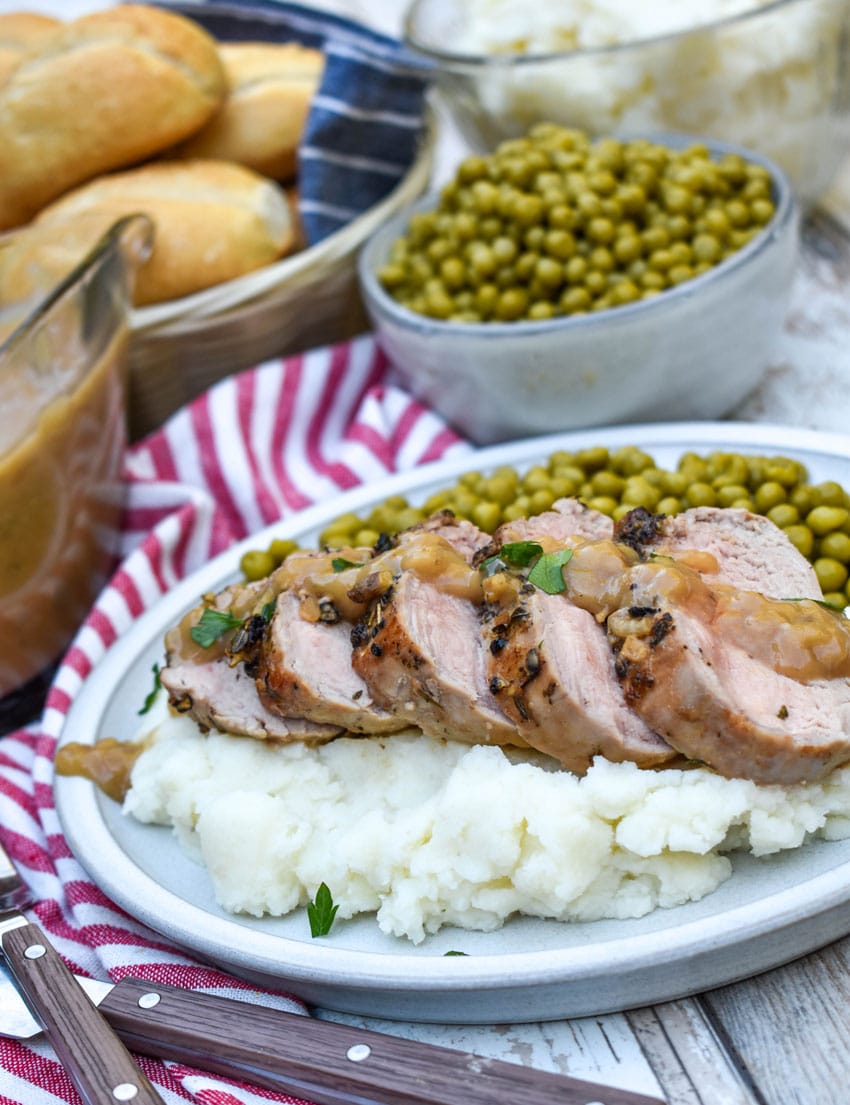 slices of pork tenderloin with apple cider gravy over mashed potatoes next to baby peas on a white plate