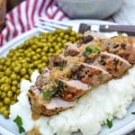 slices of pork tenderloin with apple cider gravy over mashed potatoes next to baby peas on a white plate