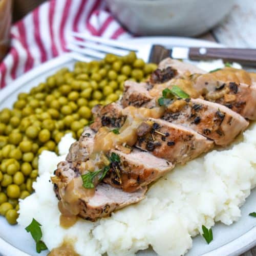 slices of pork tenderloin with apple cider gravy over mashed potatoes next to baby peas on a white plate